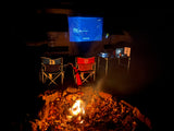 Cozy outdoor movie night next to an RV rental: campfire, folding chairs, and a projector showing "It Follows."