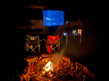 Cozy outdoor movie night next to an RV rental: campfire, folding chairs, and a projector showing "It Follows."