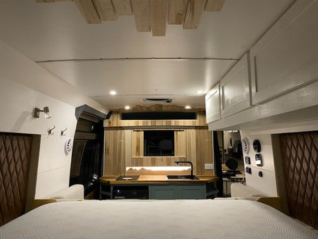A modern RV rental interior featuring a cozy bed in the foreground, a wood-paneled kitchen with a sink, and white overhead storage.