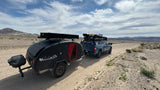 A blue RV rental truck from San Francisco Overland tows a black off-road trailer on a dusty desert road.
