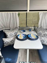 Inside an RV rental: a bright dinette area with blue seating, patterned pillows, a white table with colorful dishes.