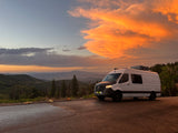 White Mercedes RV rental van at a mountain overlook. Vibrant orange sunset clouds fill the sky above scenic valleys.