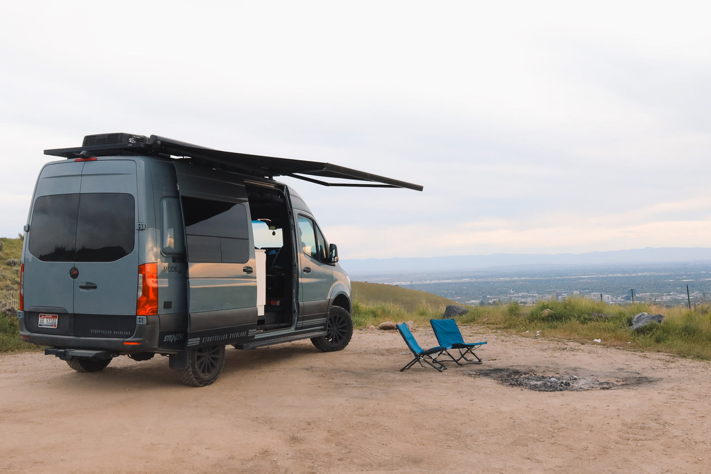 This Storyteller Overland Sprinter van is a perfect RV rental, parked on a scenic overlook with chairs and a fire pit.
