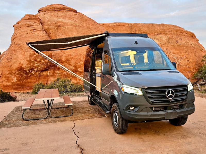 Grey Mercedes Sprinter RV rental camper van with awning & picnic table at a desert campsite with red rock formations. — photo 1