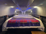 RV rental: Cozy camper bed with purple patterned blanket, bright white & blue lights, and side storage in a camper van.