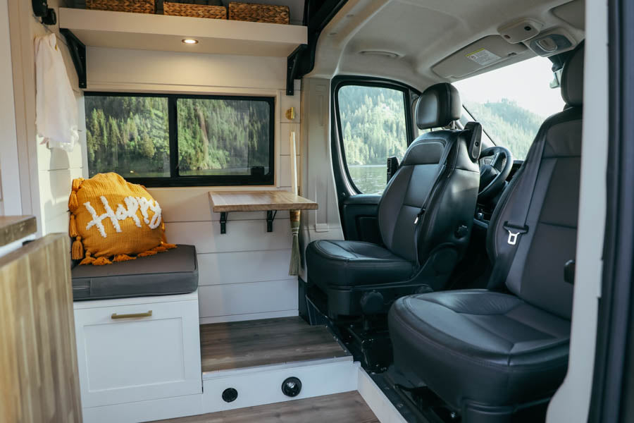 Inside a modern RV rental: a bright living space with a scenic lake view, comfy "Happy" pillow, and driver's cab.