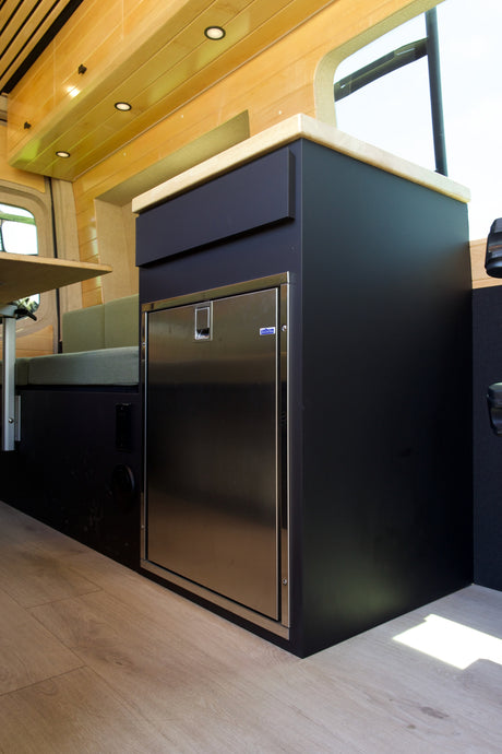 Modern RV rental interior: black kitchen cabinet, stainless fridge, wood counter, and light wood paneling with integrated lights.