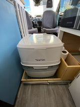 A white Porta Potti 365 portable toilet on a pull-out tray inside an RV rental, with a toilet paper holder. Ideal for van life.