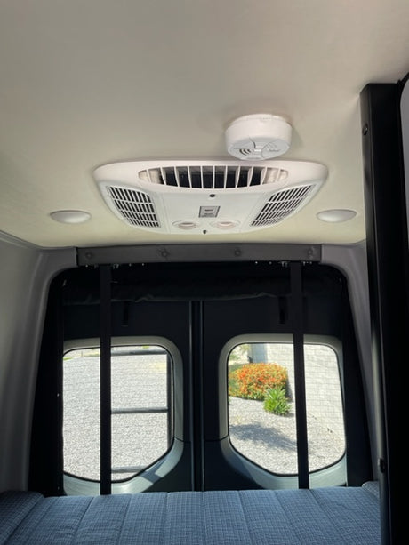 RV rental interior featuring a ceiling AC unit, wall-mounted smoke detector, and two rounded side windows.