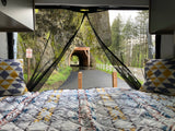 Scenic RV rental view from inside: Bed with patterned quilt, looking out to a tunnel entrance, path, and tall trees.