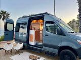 Beachside RV rental: A gray camper van with open doors reveals its cozy interior kitchen & outdoor seating. Ideal for glamping.