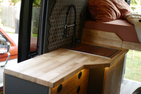 RV rental with modern camper van interior, featuring a compact kitchen with wood counters, sink, and tiled backsplash, plus a cozy bed area.