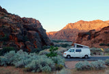 Explore stunning desert landscapes with an RV rental! A white camper van with a pop-top is parked among red rock cliffs and desert plants.