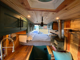 Cozy RV rental interior: bed, wood stove, kitchenette, and warm wood finishes. View out the back doors.