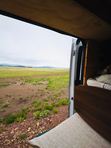 Inside an RV rental, an open door reveals a cozy bed & wooden interior, with a vast grassy field and mountains beyond.