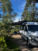 An RV rental camper van parked in a sunny forest campsite with an extended awning, surrounded by tall pine trees and blue sky.
