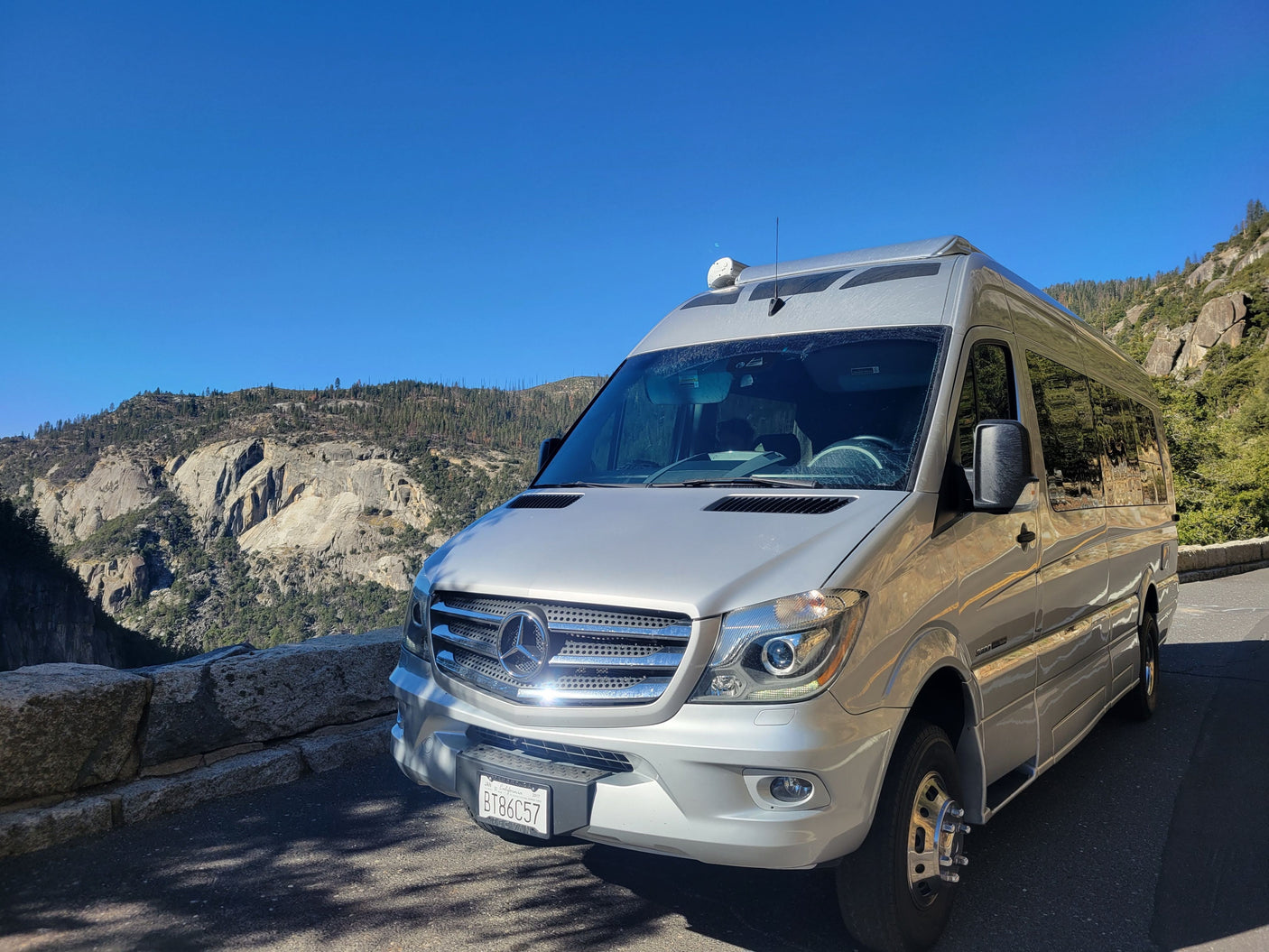 A silver Mercedes Sprinter RV rental parked at a scenic mountain overlook with rocky cliffs and a clear blue sky.