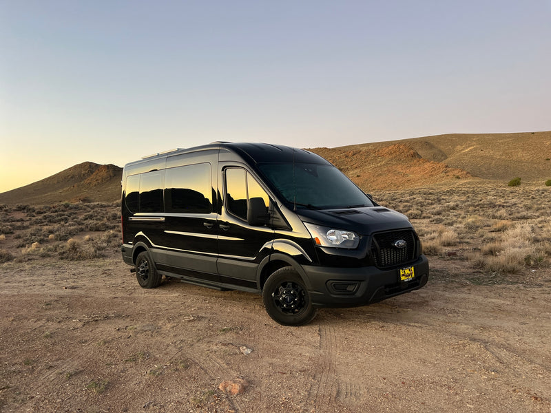 Black Ford Transit camper van RV rental parked in a desert landscape with hills at sunset, ready for adventure. — photo 7
