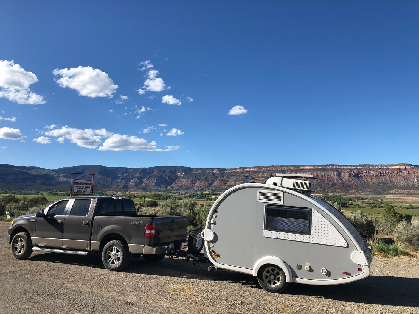 This RV rental features a grey pickup truck towing a teardrop trailer, parked by scenic mountains and green valleys under blue skies.