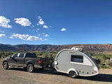 This RV rental features a grey pickup truck towing a teardrop trailer, parked by scenic mountains and green valleys under blue skies.