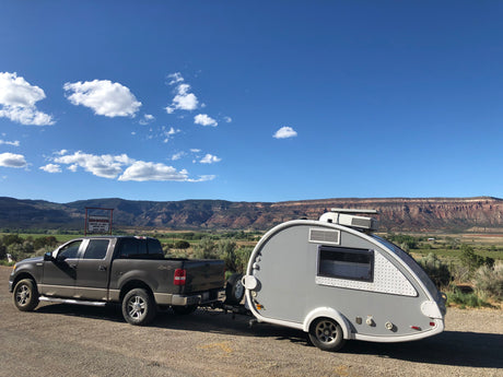 This RV rental features a grey pickup truck towing a teardrop trailer, parked by scenic mountains and green valleys under blue skies.