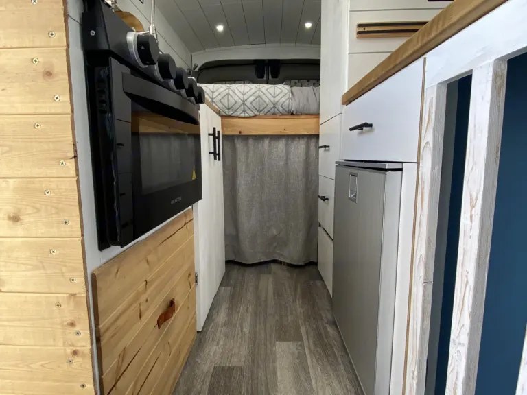Inside an RV rental: view of a compact kitchen with oven and fridge, leading to a raised bed. — photo 11