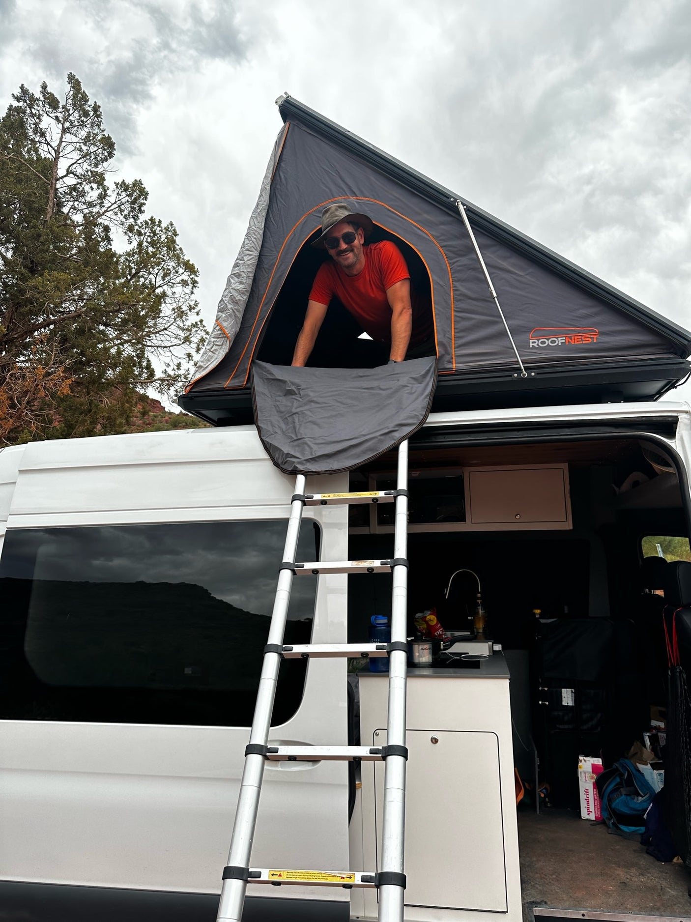 Man smiles from his RV rental's rooftop tent, accessed by a ladder from the camper van. Perfect for outdoor adventures!