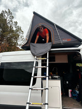 Man smiles from his RV rental's rooftop tent, accessed by a ladder from the camper van. Perfect for outdoor adventures!