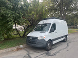 White Mercedes-Benz Sprinter van, potentially an RV rental, parked curb-side on a residential street with trees and a black roof rack.