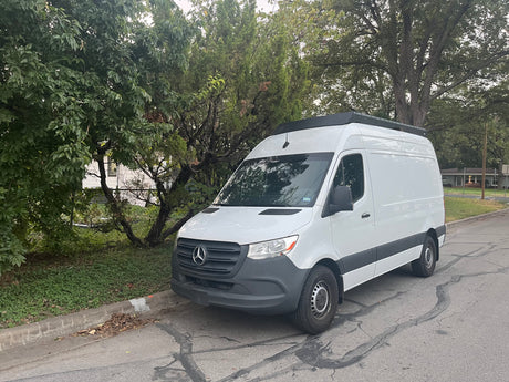 White Mercedes-Benz Sprinter van, potentially an RV rental, parked curb-side on a residential street with trees and a black roof rack.
