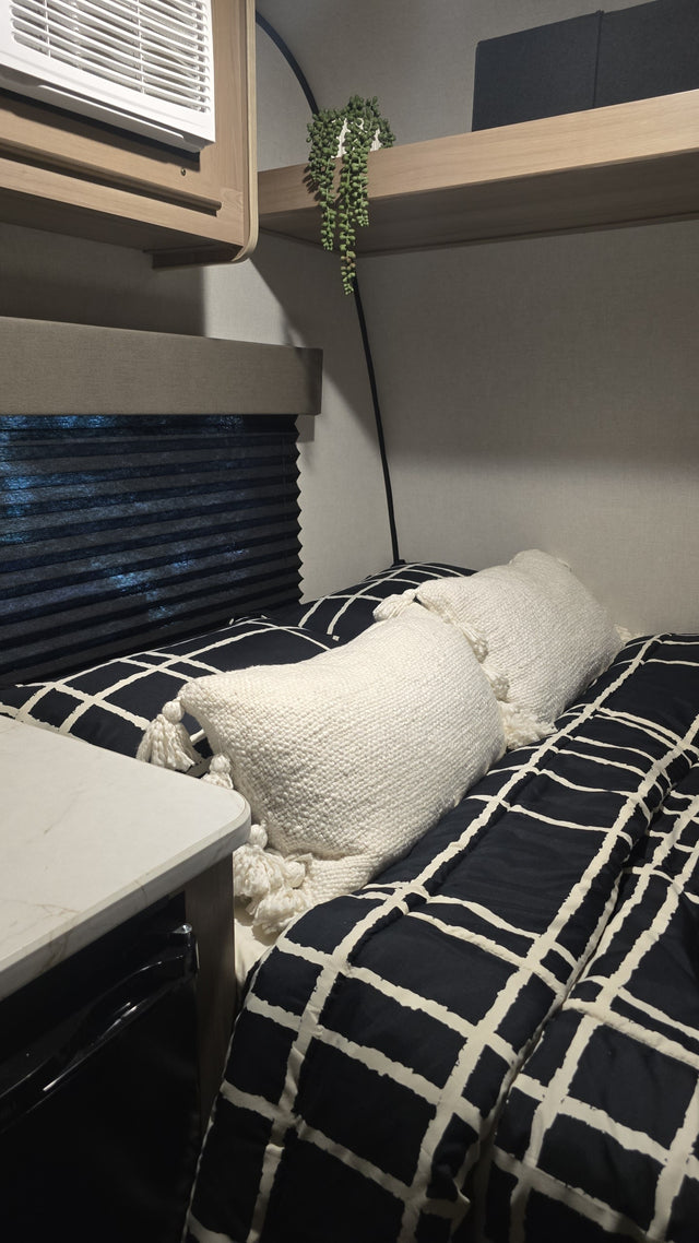 Cozy RV rental interior: A bed with black & white bedding, white pillows, window blinds, and overhead AC unit.