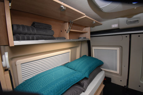 Cozy RV rental interior featuring overhead storage with towels, a comfortable bed, and windows with blinds for road trip comfort.