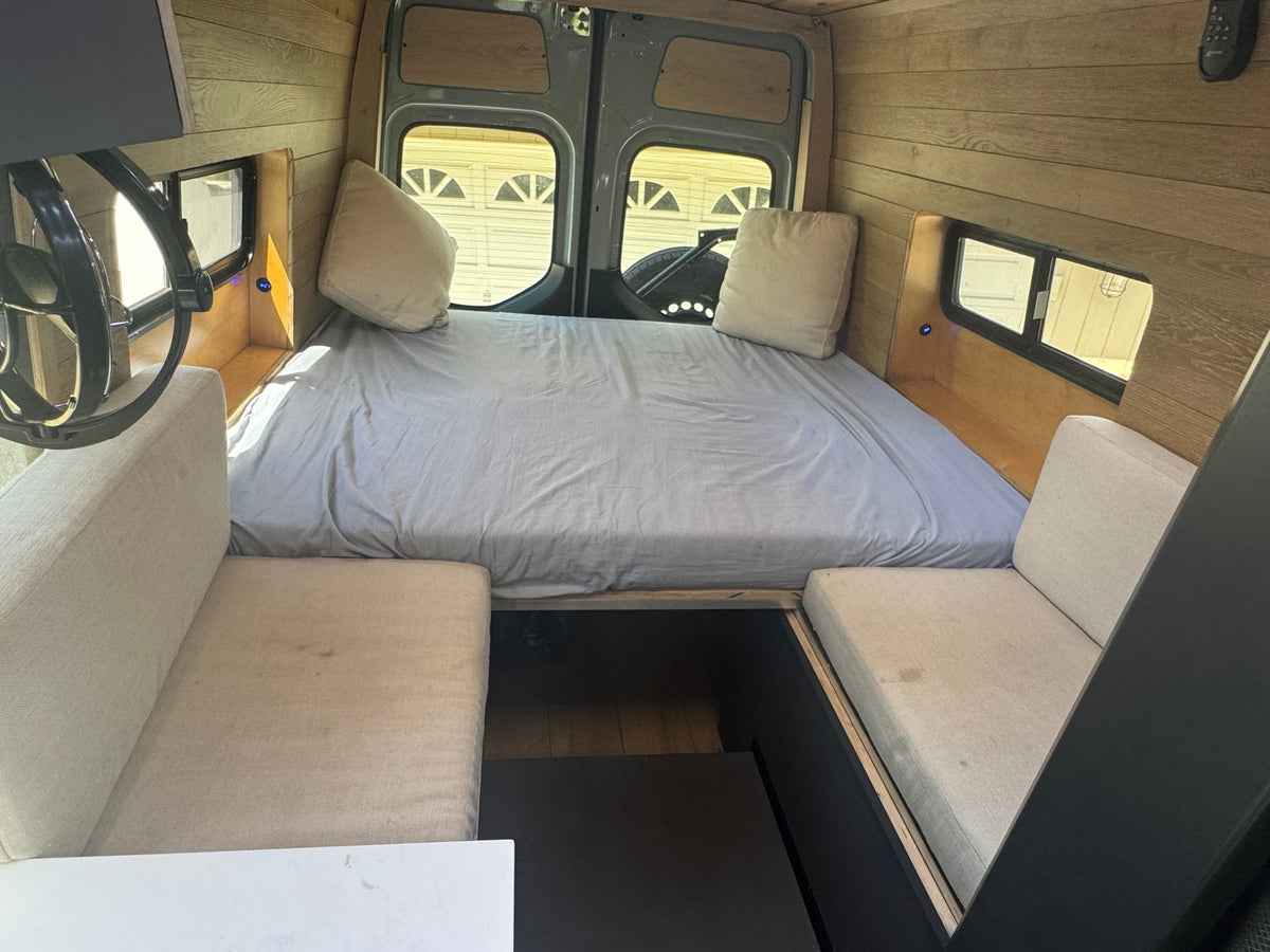 RV rental interior featuring a large bed, cozy bench seating, and warm wood finishes. Ideal for van life adventures. — photo 13