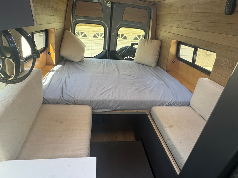 RV rental interior featuring a large bed, cozy bench seating, and warm wood finishes. Ideal for van life adventures. — photo 13