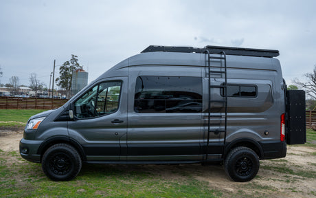 A custom gray Ford Transit camper van, perfect for RV rental adventures, with a roof rack, side ladder, and all-terrain tires.