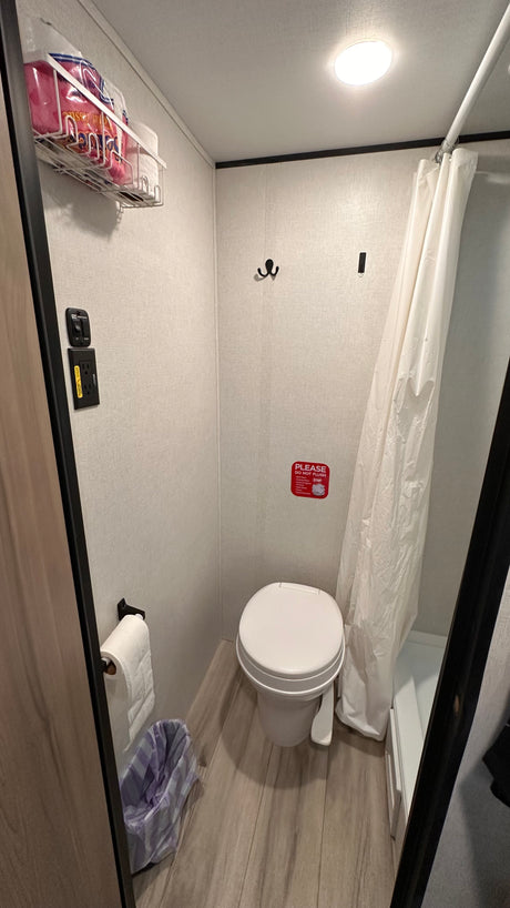 This RV rental bathroom features a toilet, shower, wall-mounted storage, light switches, and a waste bin.