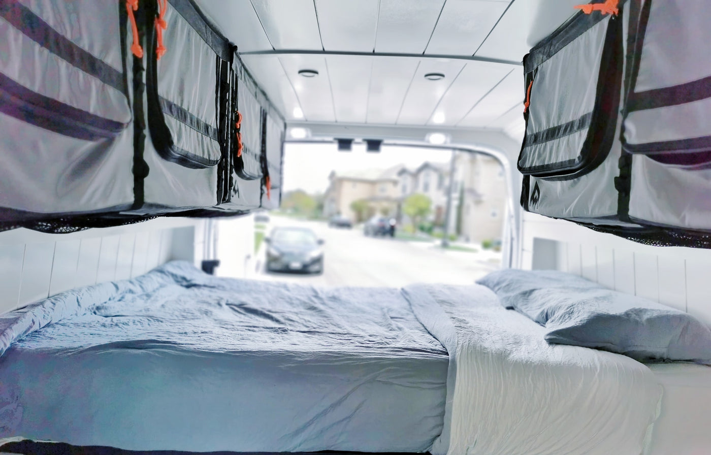 Cozy RV rental interior: bed with light blue bedding, overhead fabric storage, and a street view out back. — photo 14