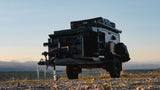 A rugged black overlanding trailer with a rooftop tent and gear, parked on a dirt path with mountains in the background. Perfect for an RV rental adventure.