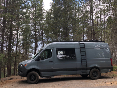 Gray Sprinter RV rental van with off-road tires and side ladder, parked in a scenic pine forest.