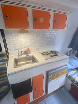 Cozy RV rental kitchen with vibrant orange cabinets, subway tile backsplash, sink, stove, and compact fridge. Perfect for road trips.