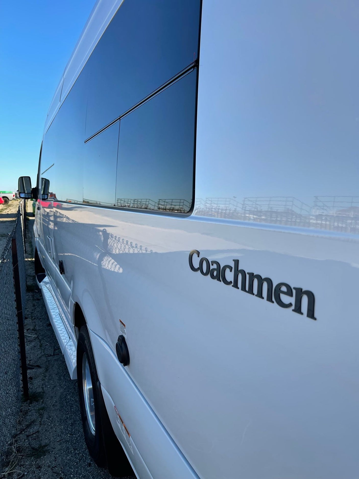 White Coachmen RV rental: side view shows tinted windows reflecting blue sky and fence.