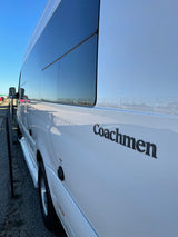 White Coachmen RV rental: side view shows tinted windows reflecting blue sky and fence.