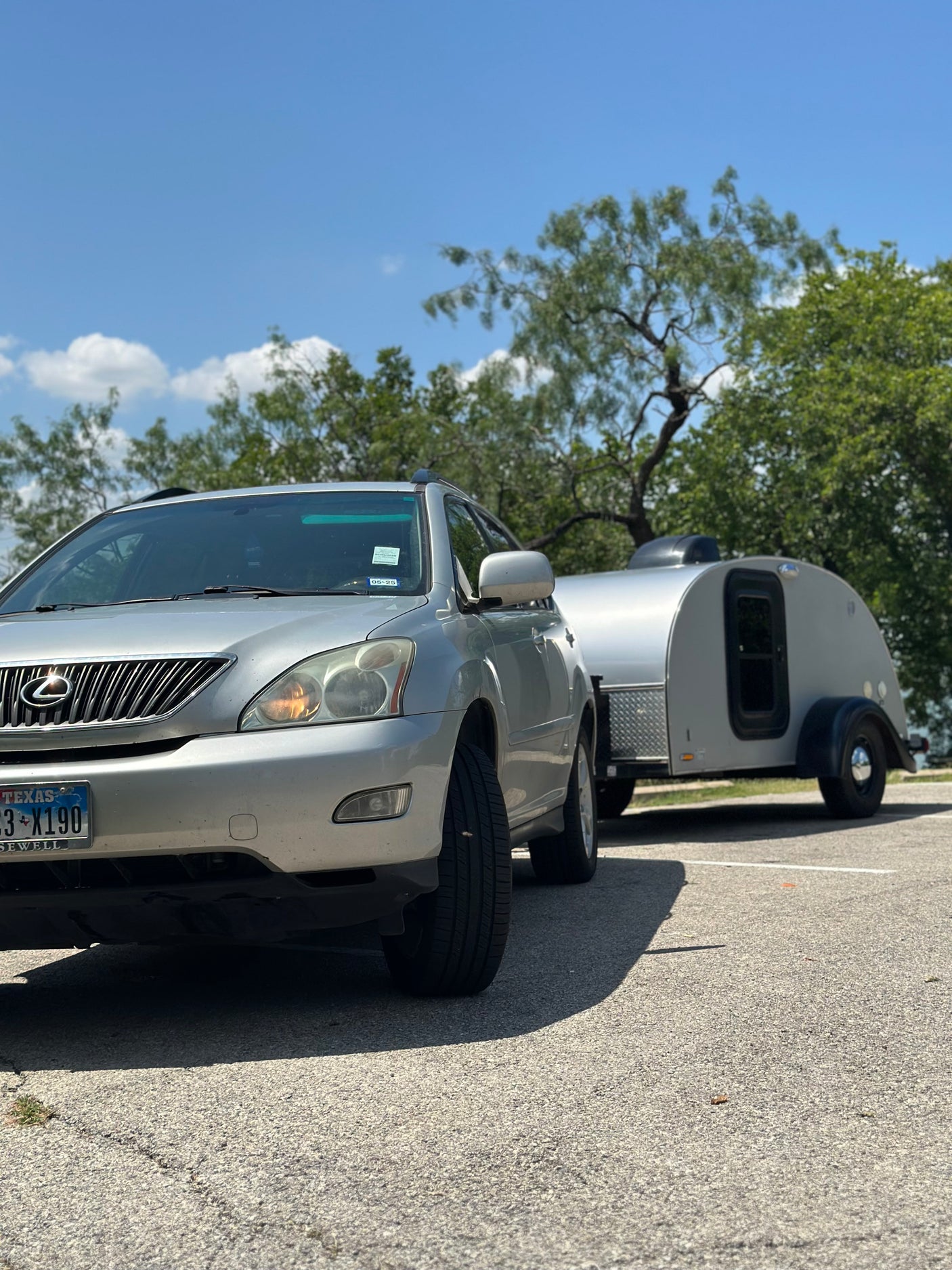 A silver Lexus SUV hitches a small, grey teardrop RV rental, parked outdoors under a blue sky.