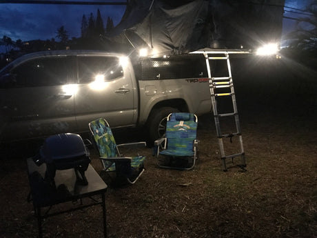 Night scene with a pickup truck, its rooftop tent and ladder set up for camping. Two chairs and a grill are ready for an RV rental trip.
