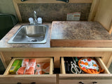 Compact RV rental kitchen with sink, countertop, and open drawers showing supplies like dishcloths and utensils.