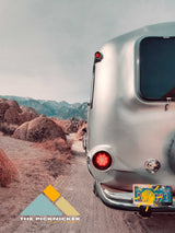 A shiny silver Airstream RV rental on a dirt road, amidst rugged desert rocks and distant mountains, ideal for adventure.