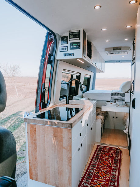 A fully equipped RV rental camper van interior. Features include a kitchen, bed, and open rear doors to scenery.