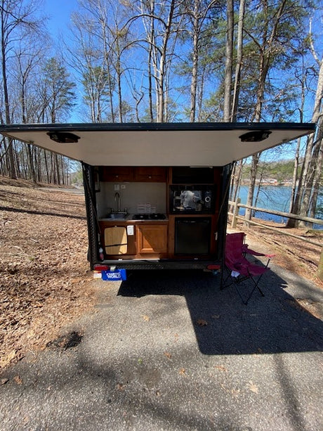 Rear of an RV rental trailer open, revealing a compact outdoor kitchenette. Parked near a lake, ready for camping.
