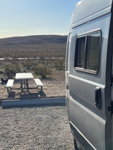 A light grey RV rental parked at a desert campsite, with a picnic table ready for outdoor dining and scenic hills in the background.