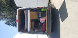 An RV rental camper van's open rear doors reveal a well-organized interior with rolled bedding, a portable fridge, and storage.